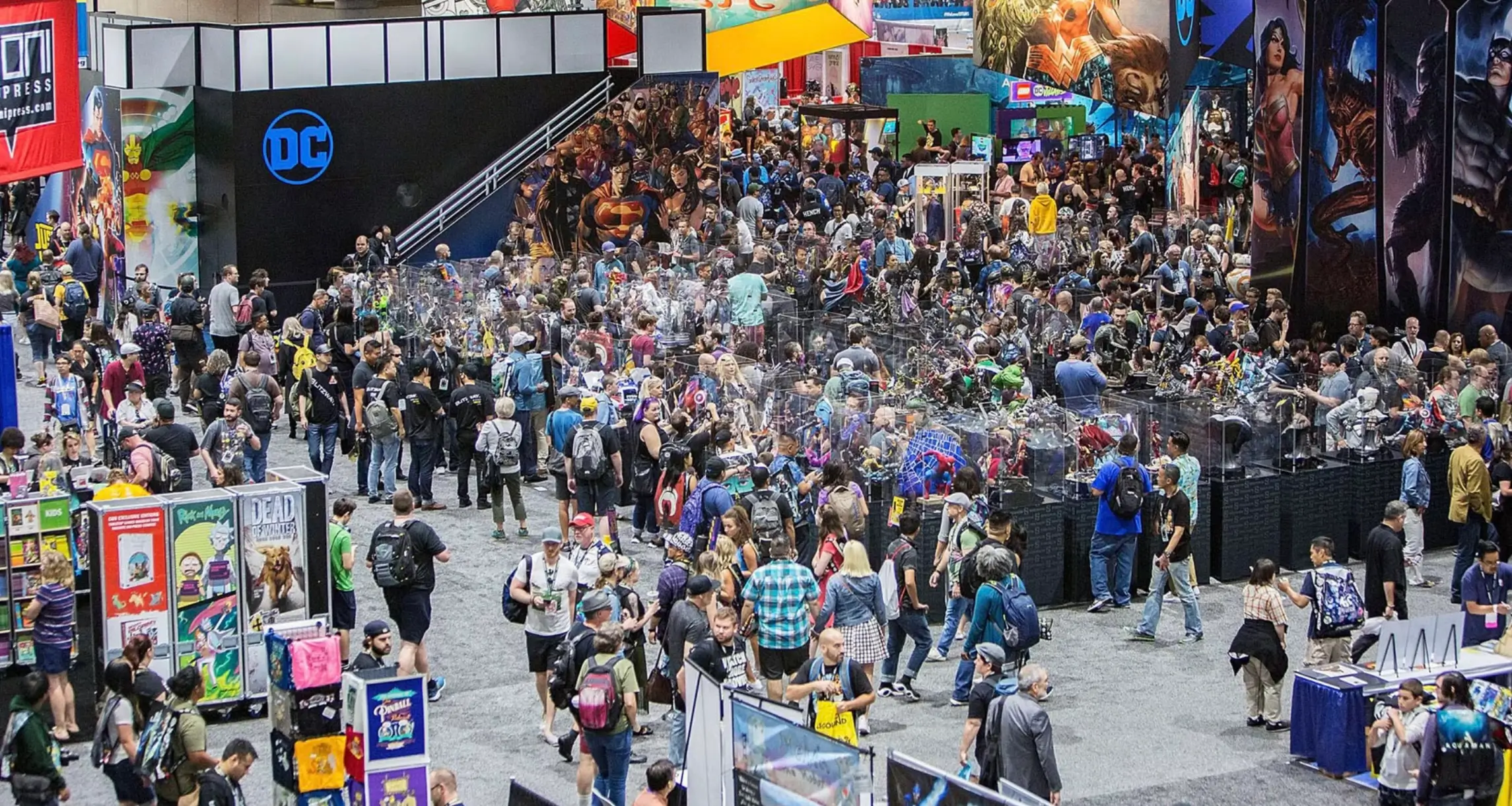 A wide overhead view of a packed convention hall during San Diego Comic-Con, with thousands of attendees moving between large publisher booths, collectible displays, artist tables, and gaming exhibits. A prominent DC booth with a glowing blue logo anchors the left side, while towering banners featuring Batman, Wonder Woman, Superman, and other comic book characters overlook the bustling show floor. Glass display cases filled with statues and collectibles sit at the center as visitors browse, photograph exhibits, and gather throughout the expansive exhibition space.