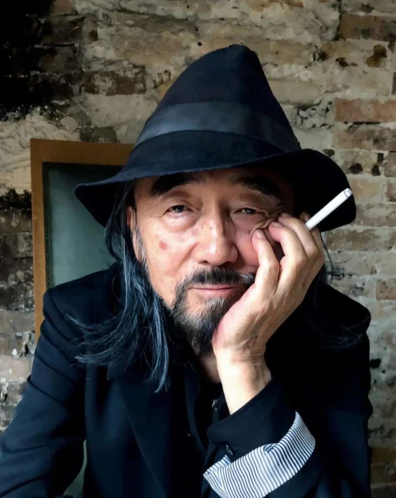 A close portrait of Yohji Yamamoto wearing a black fedora and dark blazer, resting his face on one hand while holding a cigarette. He gazes calmly toward the camera against a weathered brick wall, with long gray hair framing his face