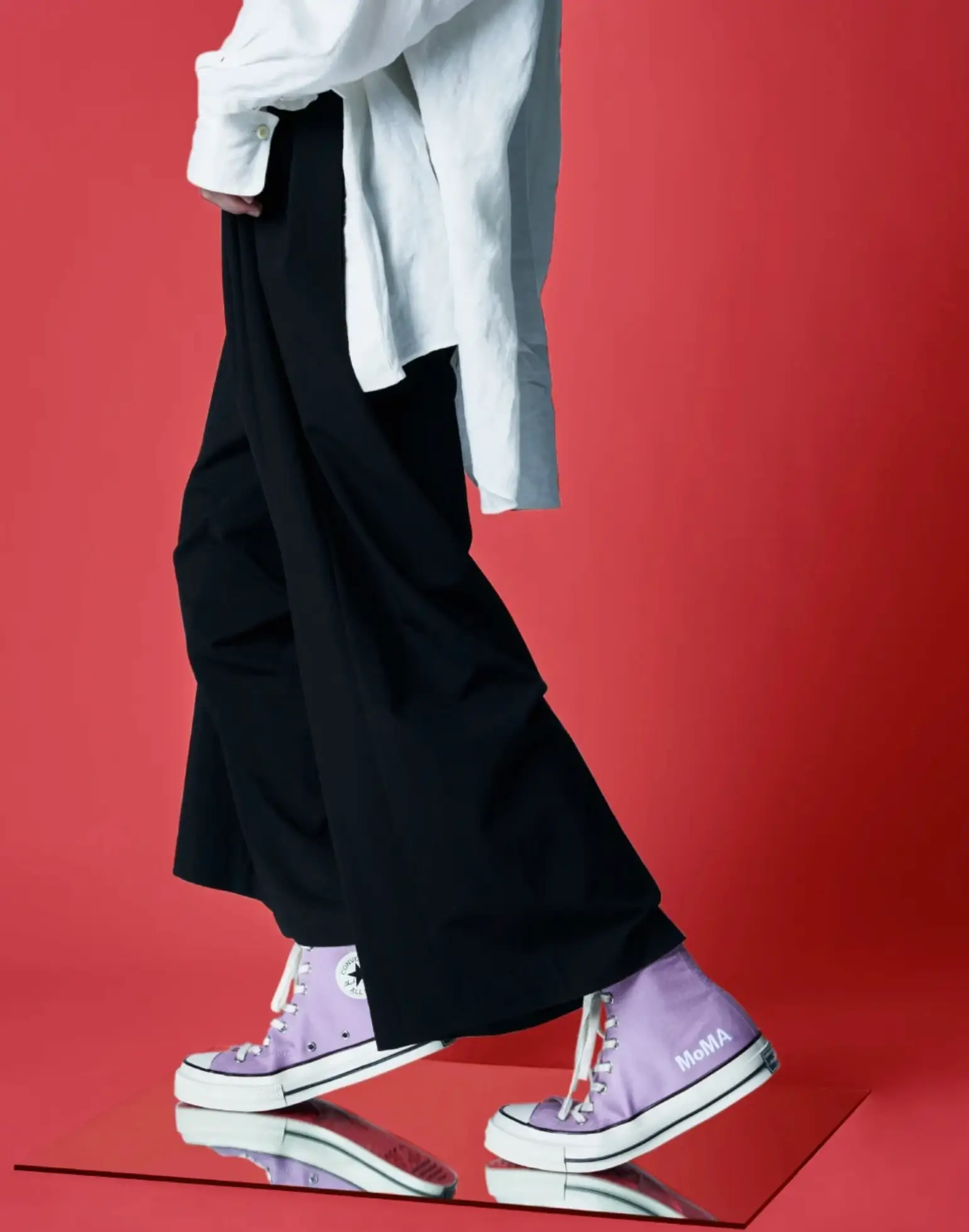 Model wearing lavender Converse Chuck Taylor All Star high-top sneakers from the MoMA collaboration, paired with wide black trousers and an oversized white shirt against a vivid red studio backdrop. The minimalist campaign image highlights the pastel canvas upper, classic rubber foxing, and MoMA branding on the heel.