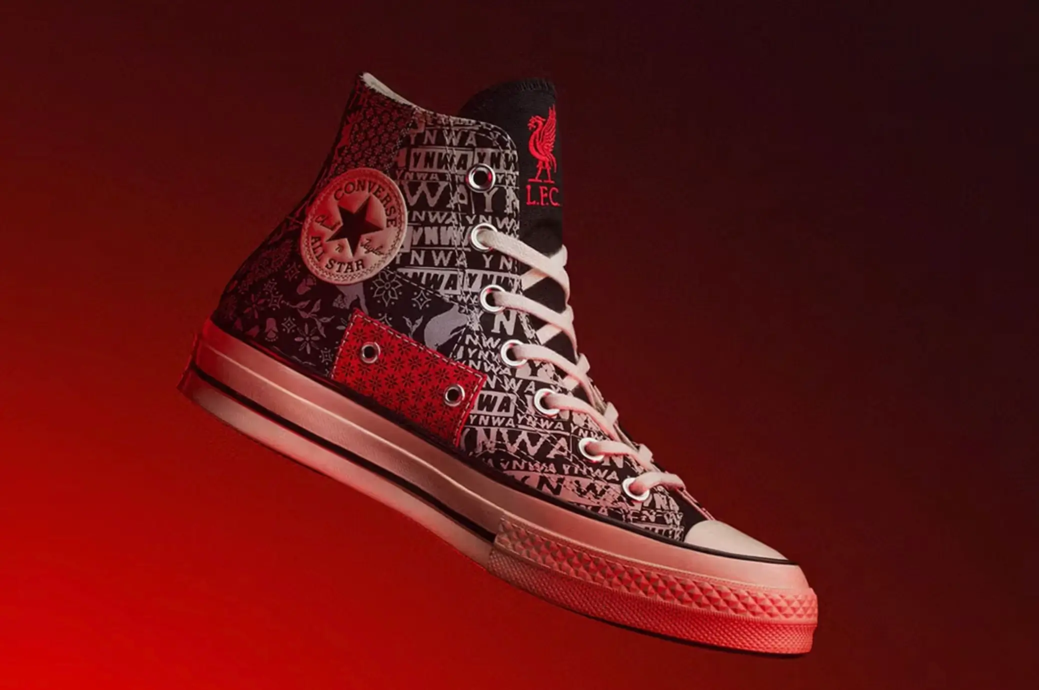 Liverpool FC × Converse Chuck Taylor All Star high-top sneaker shown in a dramatic studio campaign image against a red gradient background. The black canvas upper features YNWA typography, tonal graphic patterns, a red heel overlay, white laces, the classic Converse ankle patch, and the Liverpool club crest on the tongue.