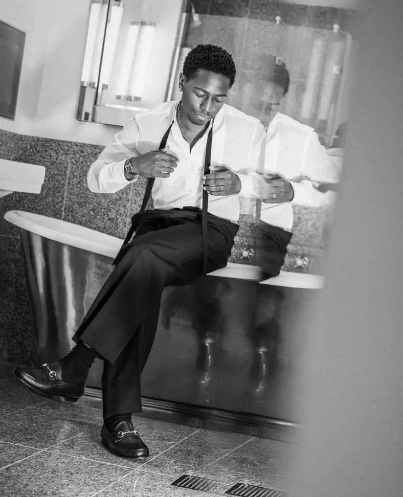 British actor David Jonsson sits on the edge of a freestanding bathtub in a monochrome portrait, adjusting his suspenders while wearing a crisp white dress shirt, tailored black trousers, and black leather loafers. Reflections in the mirror and soft natural lighting create a candid, editorial atmosphere that emphasizes classic menswear styling.