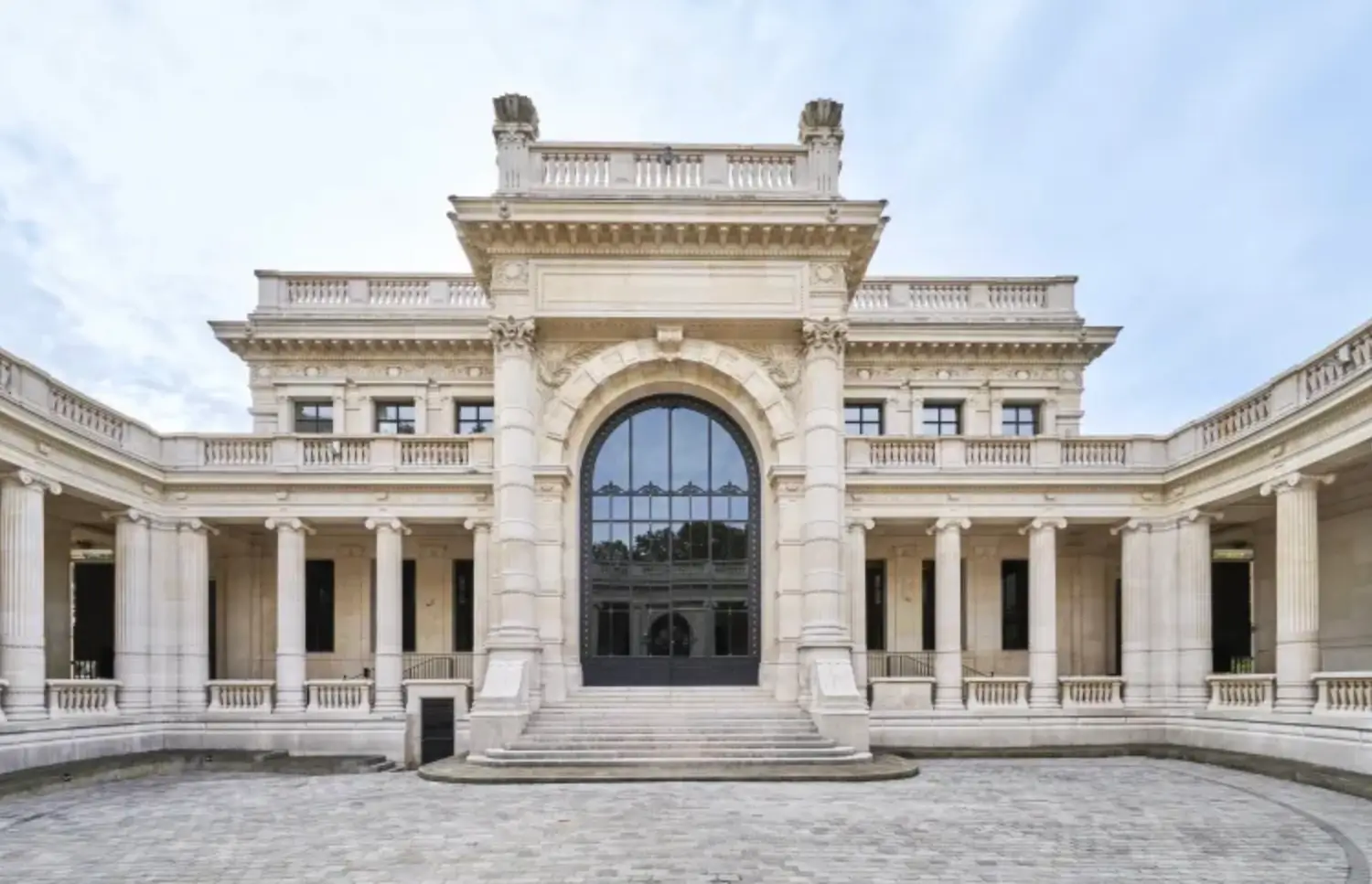 Grand neoclassical stone building with a symmetrical colonnaded façade, central arched entrance, and broad courtyard beneath a clear blue sky.