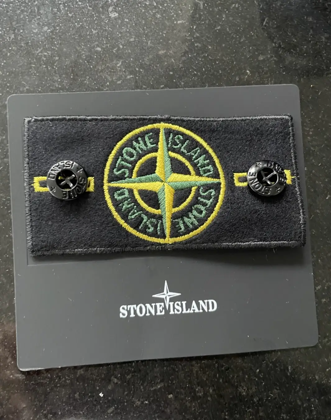 Stone Island compass badge displayed on its branded presentation card, featuring the iconic embroidered green and yellow compass logo on a black rectangular patch with two black removable logo buttons attached on either side.