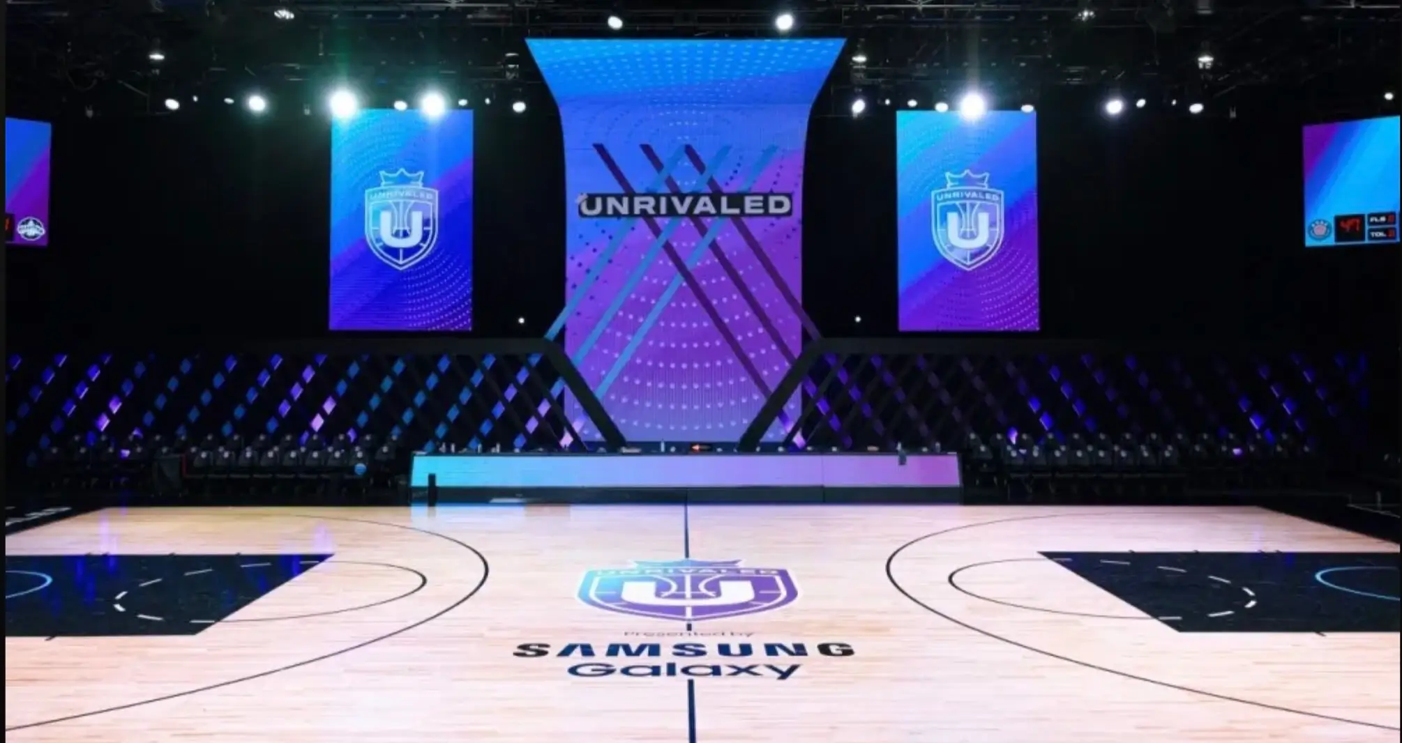 Wide view of the Unrivaled basketball arena featuring a polished hardwood court with the league's crest at center court, Samsung Galaxy branding, black seating surrounding the floor, and towering blue-and-purple LED displays showcasing the Unrivaled logo above the court.
