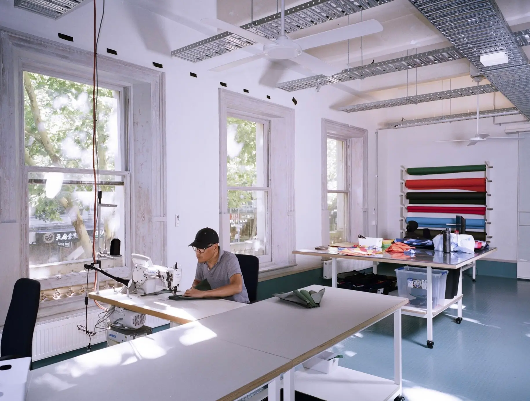 Inside the Crumpler design studio, a maker operates an industrial sewing machine while large worktables, fabric rolls, and bag prototypes fill a bright workspace illuminated by tall windows and natural light.