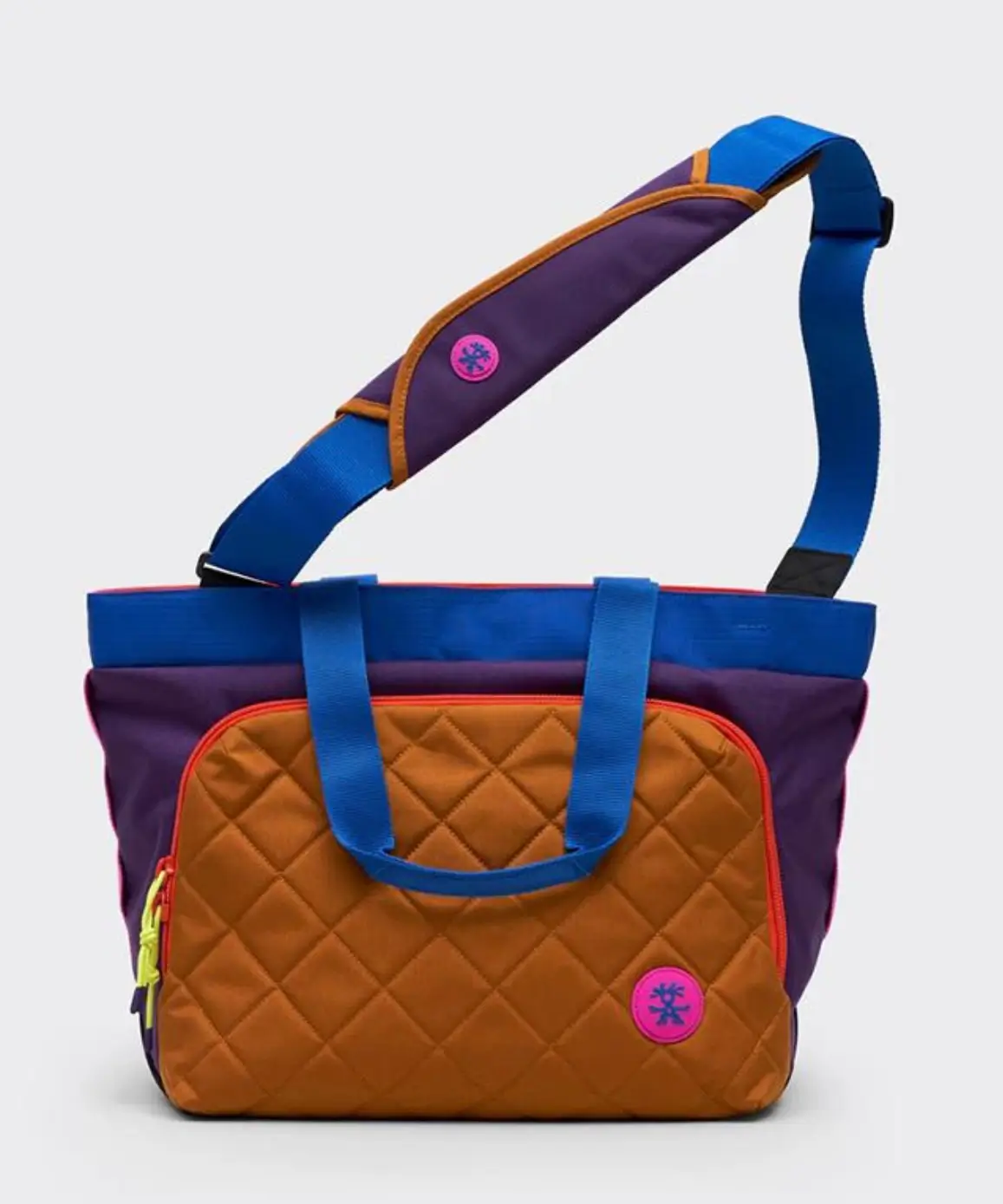 Crumpler tote bag displayed against a clean white background, featuring a quilted camel front pocket, deep purple body, bright blue carry handles and shoulder strap, neon pink logo patch, and contrasting orange trim.