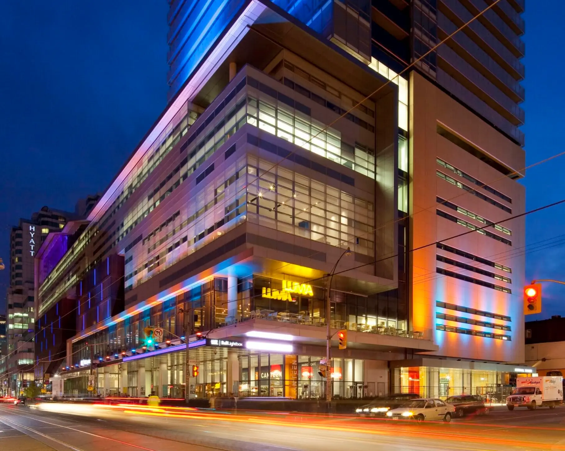Modern mixed-use high-rise building illuminated with blue, purple, and amber architectural lighting at dusk, featuring glass façades, street-level retail, and light trails from passing traffic along a busy downtown intersection.
