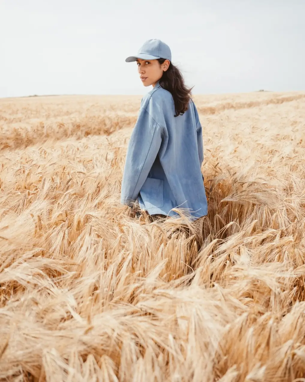 Model wearing a light blue oversized jacket and matching cap while walking through a golden wheat field