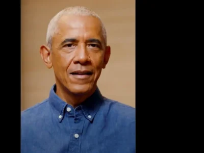Close-up portrait of Barack Obama wearing a blue button-down shirt, looking toward the camera with a neutral expression against a plain beige background