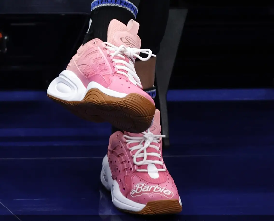 Close-up of pink Barbie-themed basketball sneakers featuring glossy pink uppers, white laces, oversized white sculpted midsoles, gum rubber outsoles, and Barbie script branding across the toe while worn with black socks on a dark blue court.