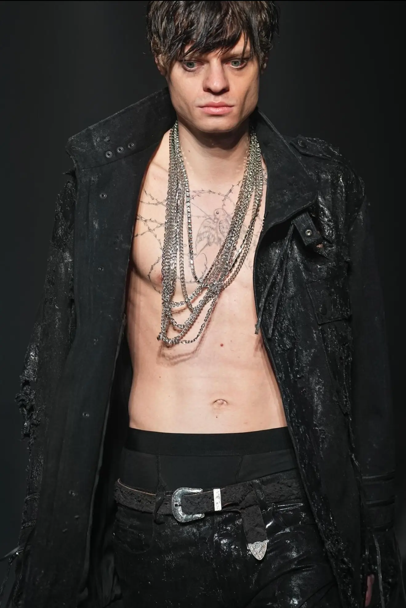 Close-up runway portrait of a model wearing a distressed black textured coat over a bare chest, layered oxidized silver chain necklaces, black leather trousers with a worn belt, and subtle chest tattoos against a dark backdrop.