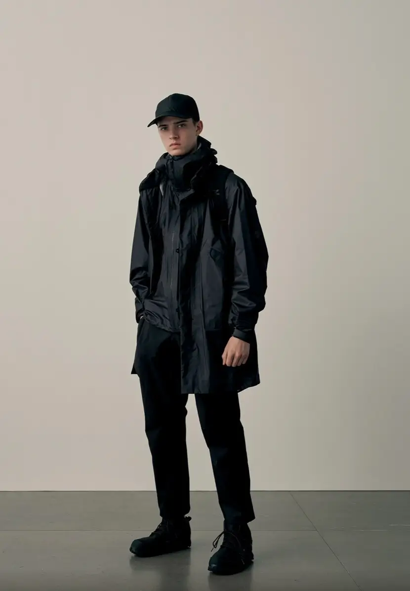 Model wearing a black DESCENTE ALLTERRAIN 81 waterproof hooded coat with matching cap, backpack, technical pants, and outdoor footwear