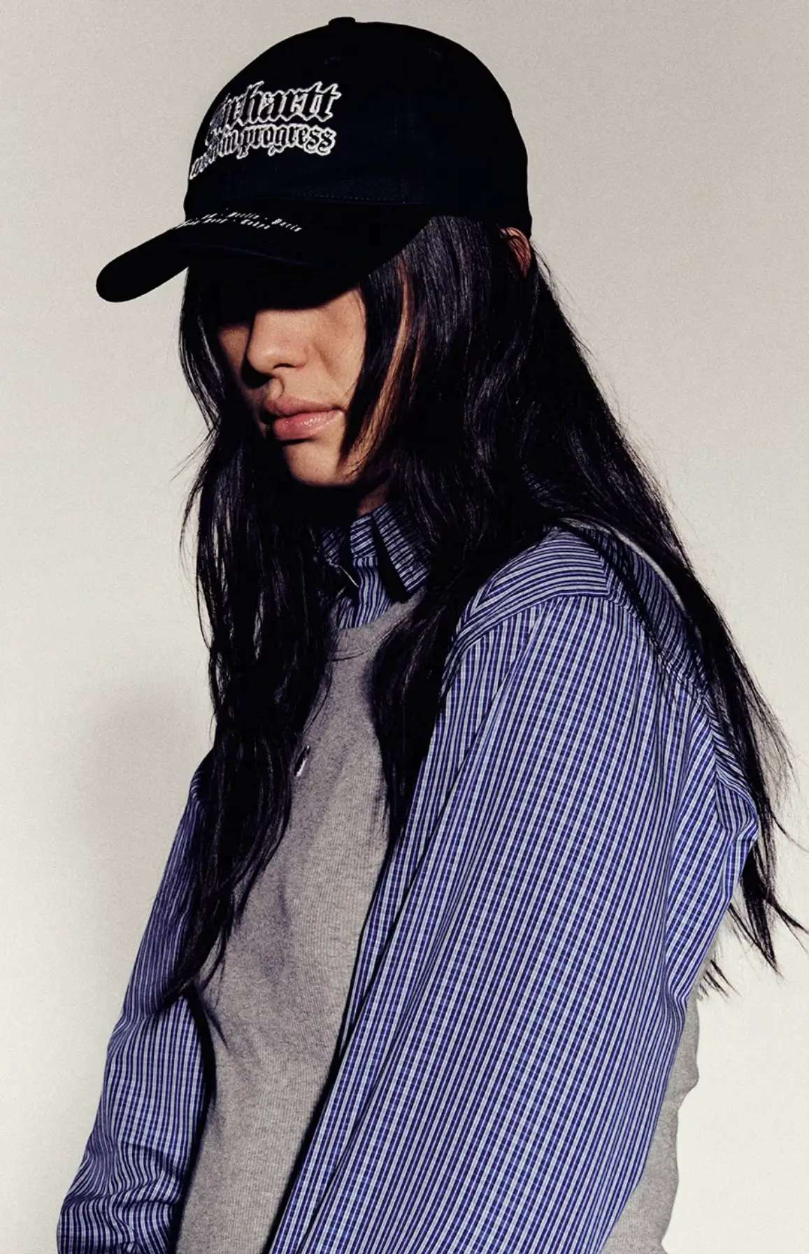 Portrait of a model wearing a black Carhartt WIP baseball cap with embroidered script logo, layered blue striped button-up shirt, and gray knit vest, showcasing minimalist contemporary streetwear styling against a neutral studio backdrop.