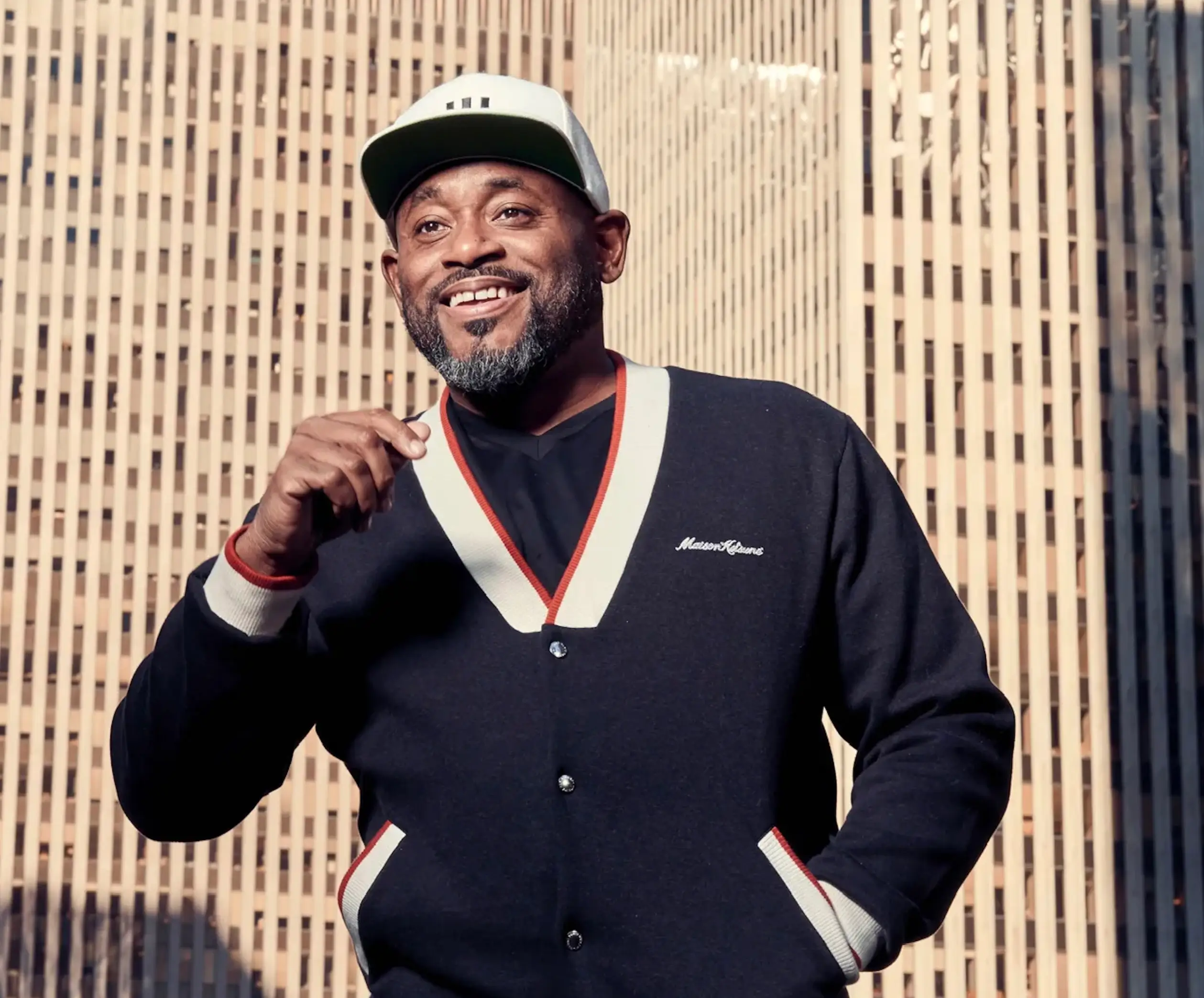 Steve Stoute smiling outdoors in front of New York City skyscrapers, wearing a white baseball cap, black V-neck cardigan with contrasting trim, and a black shirt while posing in natural sunlight.
