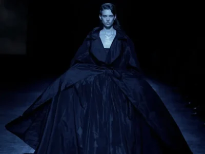 Model walking the runway in a dramatic midnight-black oversized coat dress with a voluminous floor-length skirt, wide sculptural lapels, and a tied waist, presented under moody spotlight lighting against a dark runway backdrop