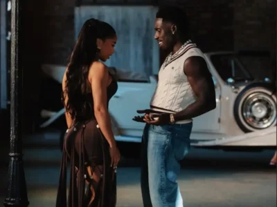 Frenna in a cream cable-knit sweater vest, light blue jeans, layered chain necklaces, and a wristwatch smiles while talking with a woman wearing a black backless dress with flowing ribbon-like panels and a high ponytail. They stand face-to-face inside an industrial warehouse or garage, with a white vintage luxury convertible parked behind them. Warm cinematic lighting, exposed brick walls, and a relaxed, intimate atmosphere give the scene a stylish editorial feel