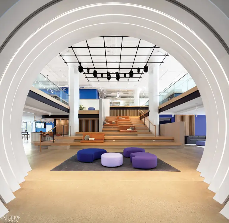 Contemporary office interior viewed through a large illuminated archway, featuring tiered wooden seating, open collaborative workspaces, and colorful circular lounge cushions.