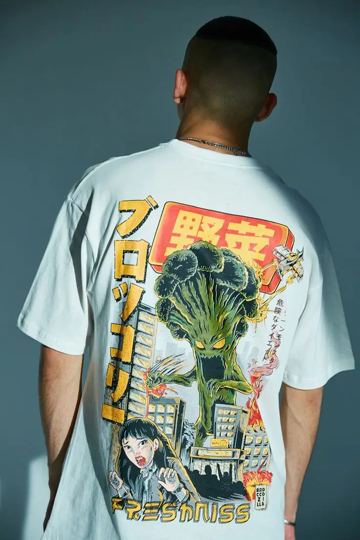 Model wearing a white oversized graphic T-shirt featuring a retro Japanese monster movie-inspired illustration of a giant broccoli creature towering over a city with manga-style artwork, bold Japanese lettering, and colorful comic graphics across the back.