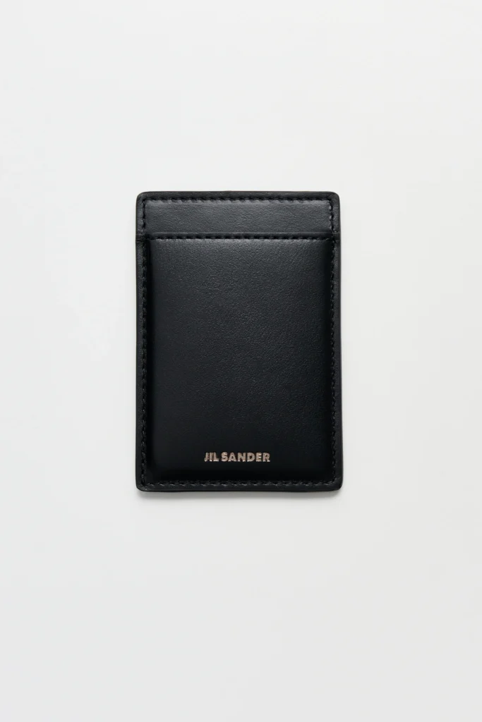 Black Jil Sander leather card holder shown from the reverse side on a clean white background. The minimalist design features a single card slot, smooth leather construction, tonal stitching around the edges, and a silver foil Jil Sander logo centered near the bottom