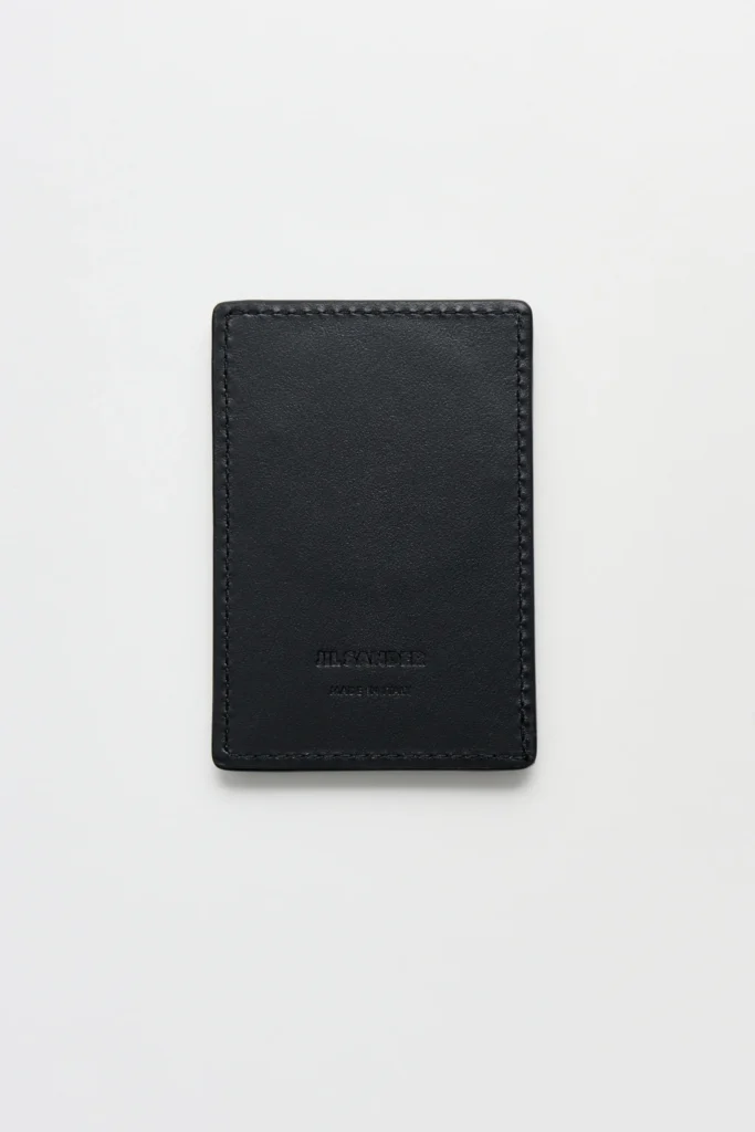 Black Jil Sander leather card holder photographed from the front on a clean white background. The minimalist card holder features smooth leather, tonal stitching around the edges, rounded corners, and a subtle embossed Jil Sander logo with "Made in Italy" centered near the bottom
