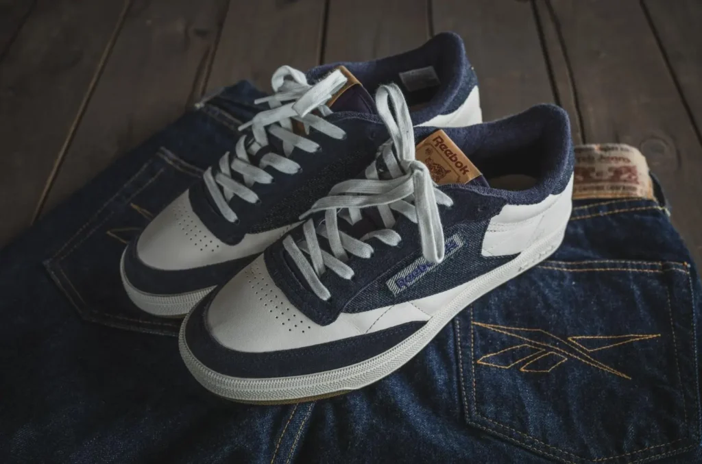 Angled view of the Reebok x TCB Jeans shoe placed across folded selvedge denim jeans on a wooden floor. White leather panels contrast with indigo denim uppers, while embroidered Reebok branding, tan tongue labels, and detailed yellow stitching on the jeans highlight the