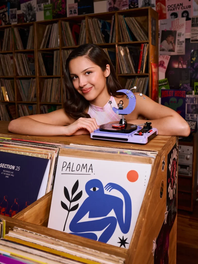 Olivia Rodrigo stands smiling in a pink plaid dress, holding a colorful LEGO flower bouquet. She poses in a vibrant record store filled with posters, vinyl records, and wooden crate