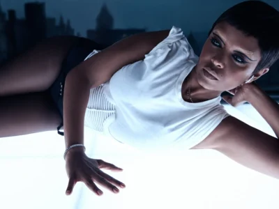Fashion portrait of a model reclining across a glowing illuminated surface at night, wearing a structured white sleeveless top with ribbed side detailing and black bottoms. A sharp pixie haircut, dramatic winged eyeliner, bleached brows, and silver jewelry complement the futuristic editorial styling, with a blurred city skyline in the background