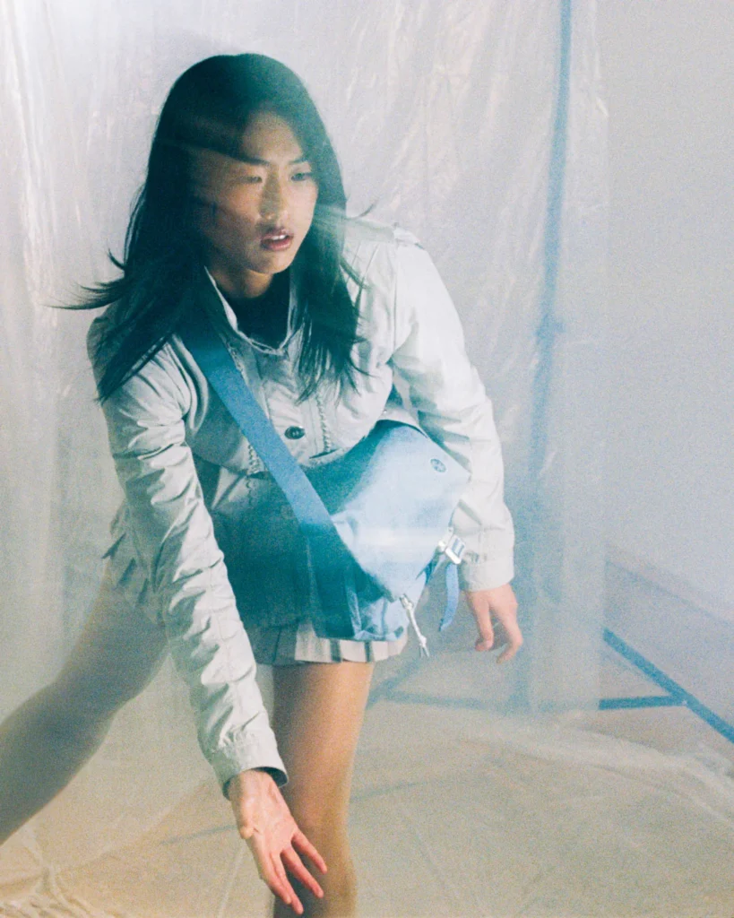 Model wearing a light blue Crumpler crossbody messenger bag in a softly lit studio with translucent plastic curtains, highlighting the bag's lightweight design and adjustable strap