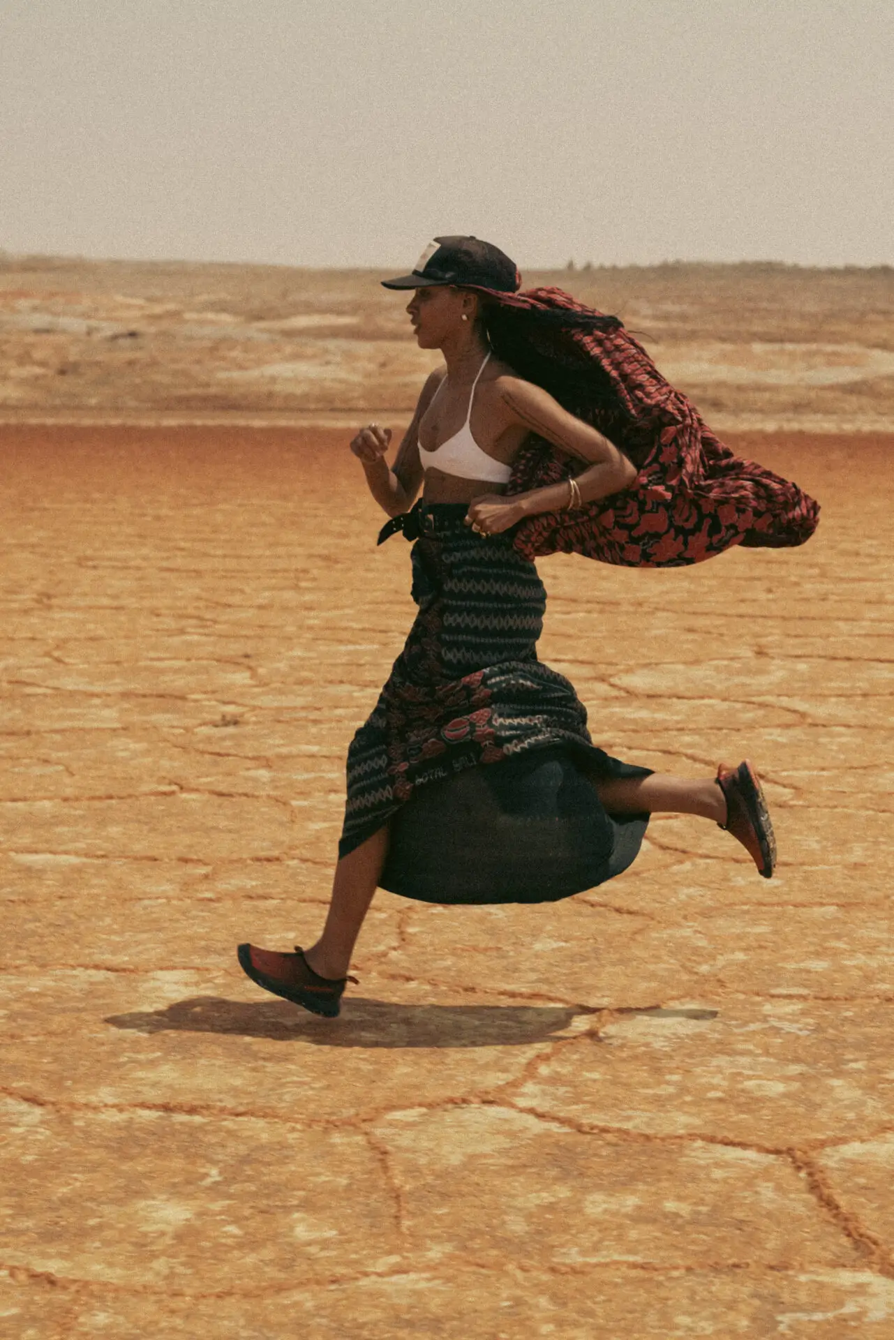 Fashion model runs across a cracked desert landscape wearing a white bralette, patterned wrap skirt, flowing red scarf, baseball cap, and leather flats in a windswept editorial scene.