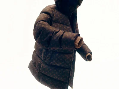 Oversized brown Louis Vuitton monogram puffer coat with an attached hood floats against a clean white background, showcasing exaggerated quilting, ribbed knit cuffs, and a partially unzipped front in a minimalist editorial product image