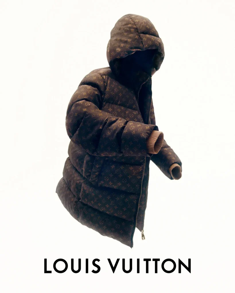 Oversized brown Louis Vuitton monogram puffer coat with an attached hood floats against a clean white background, showcasing exaggerated quilting, ribbed knit cuffs, and a partially unzipped front in a minimalist editorial product image