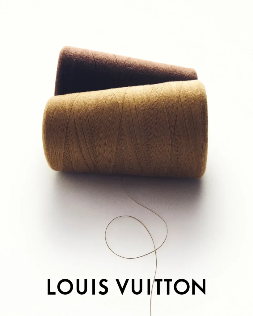 Two large spools of thread in camel and dark brown sit stacked together with a loose strand curling across the white surface, representing the craftsmanship behind Louis Vuitton's Silk Tech concept