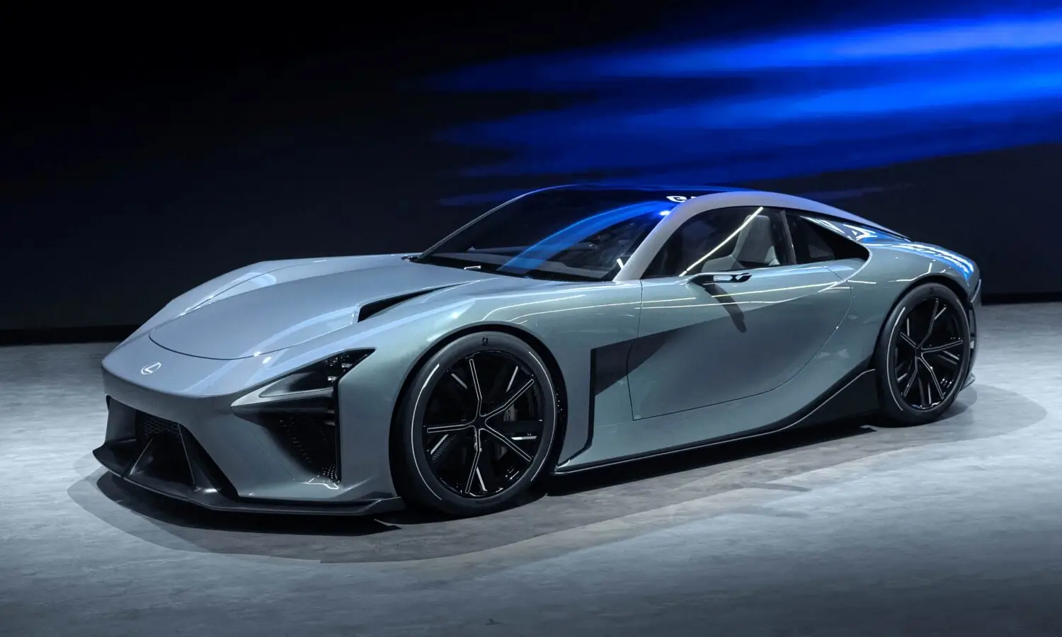 Side profile of the Lexus Electrified Sport concept in metallic silver, showcasing its low-slung supercar proportions, sculpted bodywork, black multi-spoke wheels, sharp LED lighting, and aerodynamic silhouette under dramatic blue studio lighting.