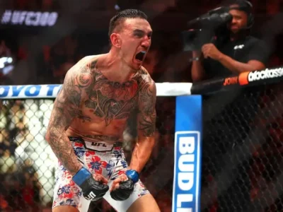 Max Holloway inside the UFC Octagon lets out a roar after a victory, wearing white floral fight shorts, blue gloves, and a blue mouthguard while displaying extensive tattoos across his chest and arms
