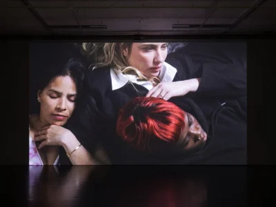 Large-scale video projection inside a dark gallery space showing three people resting closely together, creating an intimate portrait of vulnerability and human connection, with the illuminated screen reflected across the polished floor