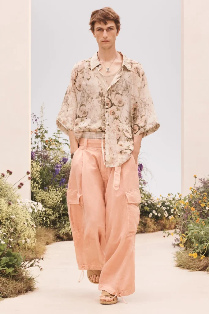 Model walking a garden runway in a relaxed floral-print short-sleeve shirt, wide blush pink cargo trousers, layered necklace, and beige sandal