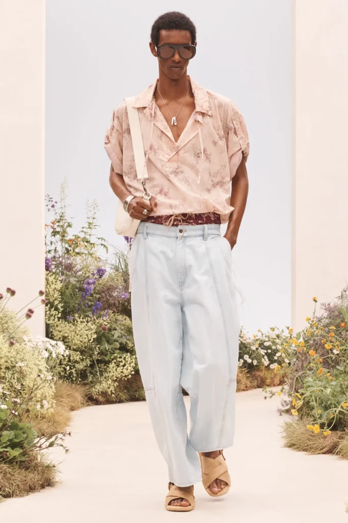 Model wearing a pale floral camp-collar shirt with oversized light blue trousers, oversized sunglasses, cream shoulder bag, and beige sandals on a floral runway