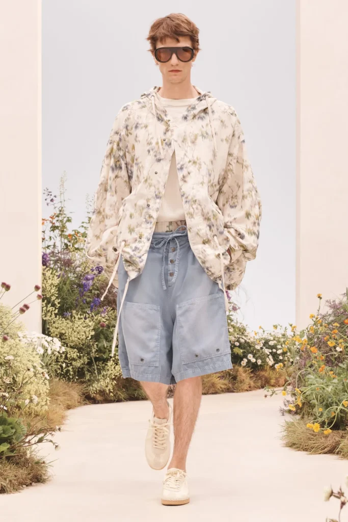 Model in a floral hooded lightweight jacket, white T-shirt, oversized light blue shorts, oversized sunglasses, and cream sneakers walking a garden runway