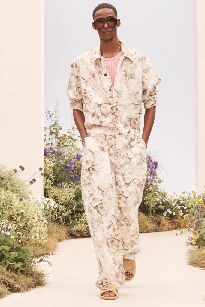 Model walking in a matching cream floral-print overshirt and wide-leg trousers layered over a pastel pink knit tank with oversized sunglasses and beige sandal