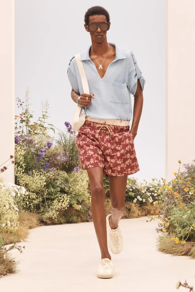 Model wearing a light blue knit polo, burgundy floral shorts, oversized sunglasses, cream shoulder bag, and white shoe on a garden runway