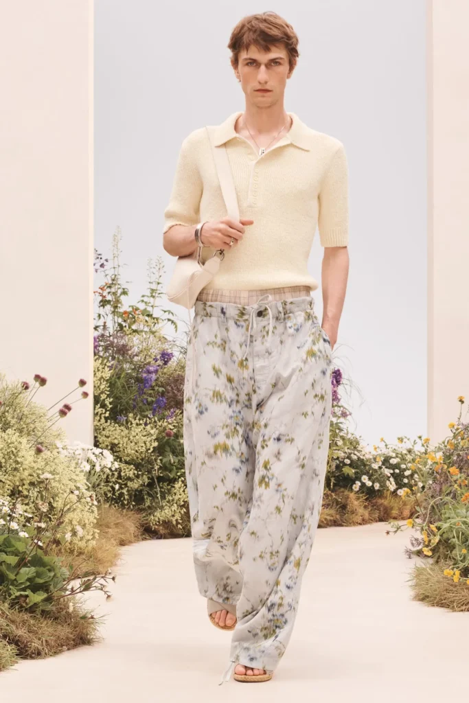 Model in a cream knit polo with loose floral-print trousers, cream shoulder bag, layered necklace, and beige sandals walking a floral runway