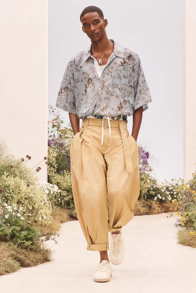 Model wearing a blue floral-print short-sleeve shirt, oversized tan pleated trousers, layered necklace, and white shoe on a flower-lined runway