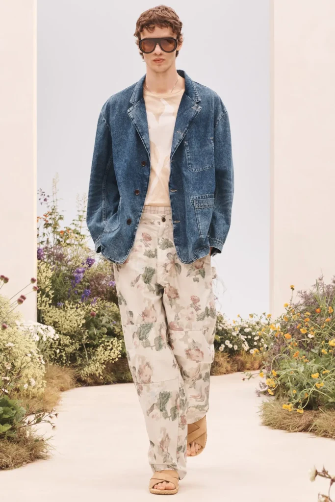 Model in a dark blue denim chore jacket, beige T-shirt, floral-print wide-leg trousers, oversized sunglasses, and beige sandals walking a garden runway