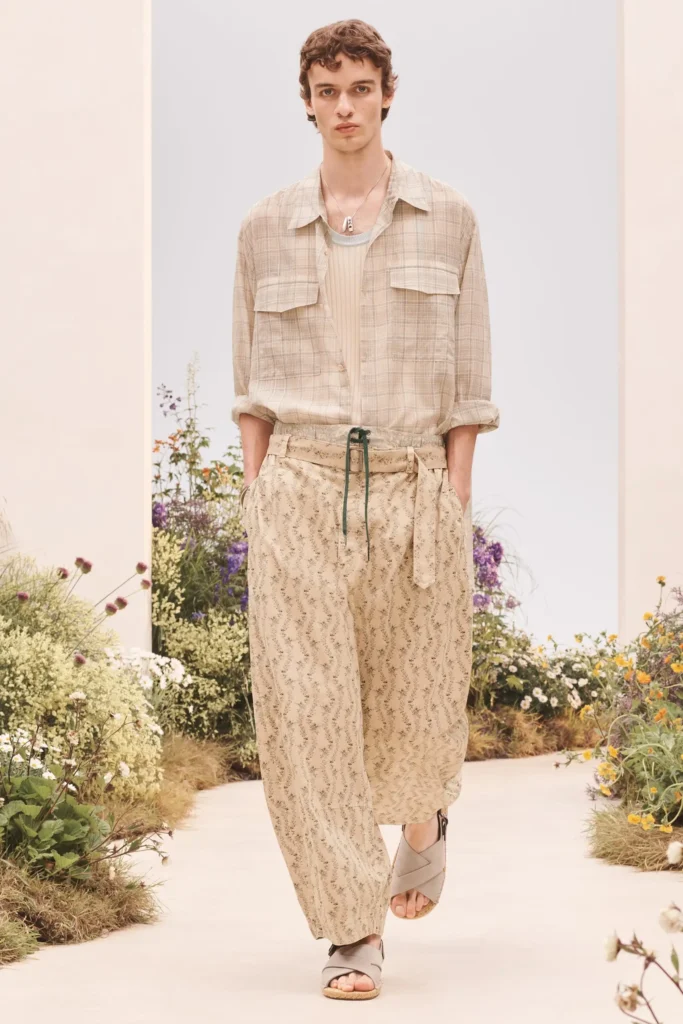 Model wearing a beige plaid overshirt layered over a ribbed knit top with loose patterned trousers, wrap sandals, and a pendant necklace on a floral runway