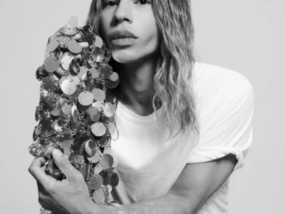 Black-and-white portrait of OLIVIER ROUSTEING with shoulder-length wavy hair wearing a plain white T-shirt, seated at a wooden table and looking directly into the camera