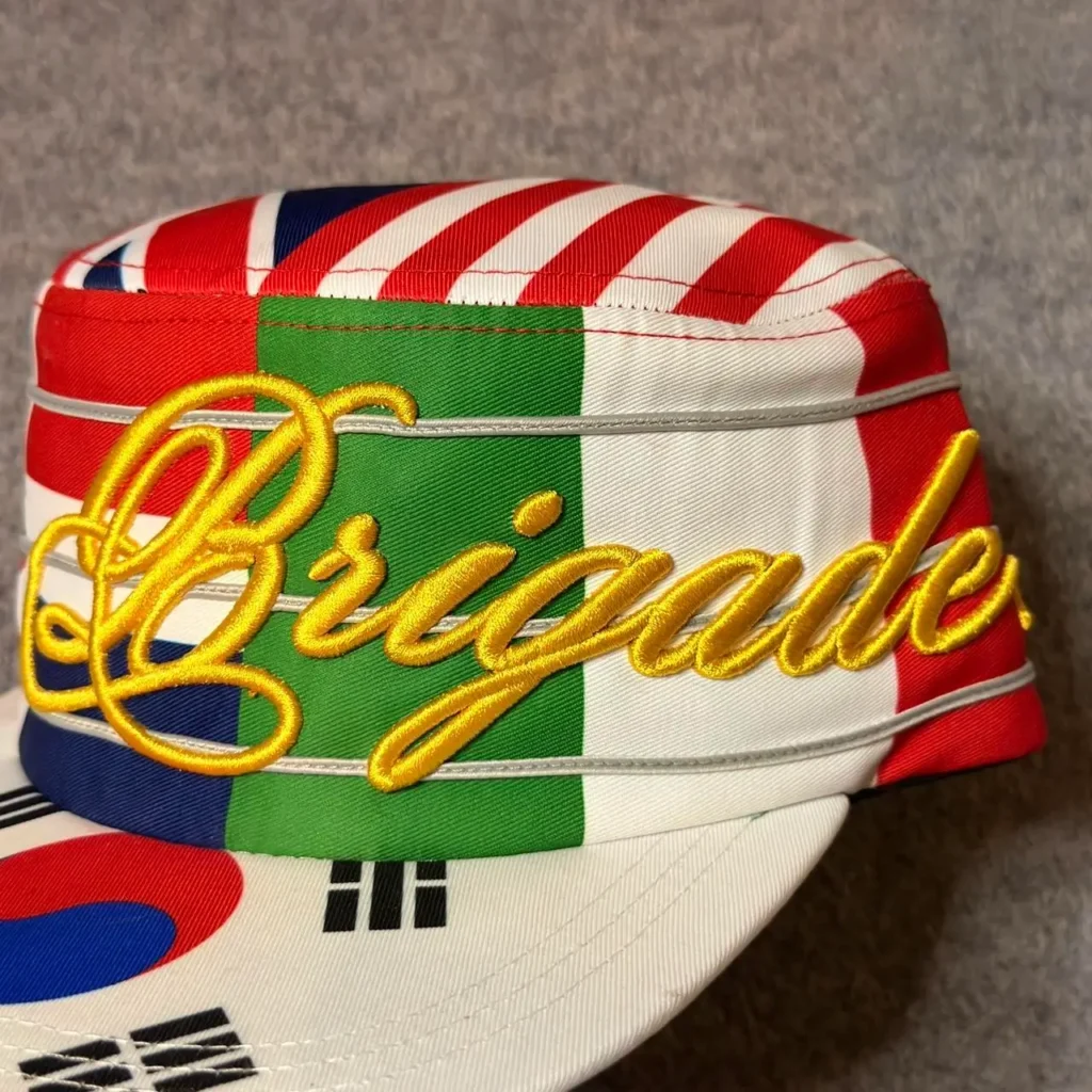 Close-up of a structured five-panel cap wrapped in a patchwork of international flag graphics, including Italy, the United States, South Korea, and Japan. Raised yellow script embroidery reading "Brigade" spans the front panel, while thin white piping accents the seams. The colorful design combines national flag motifs with bold streetwear-inspired brand