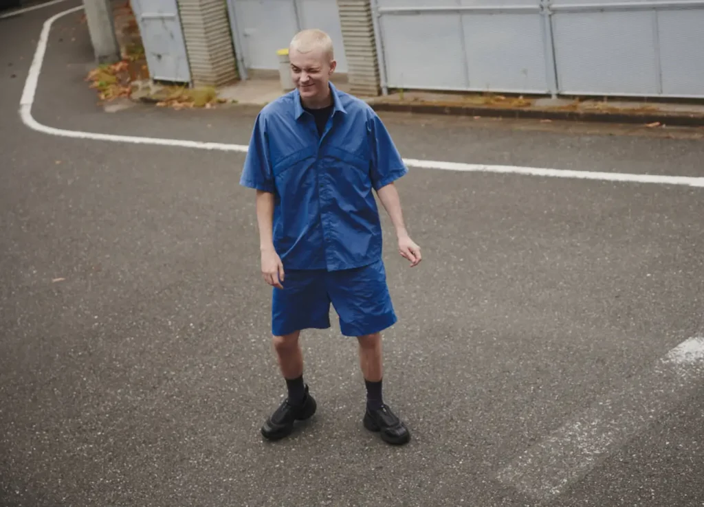 A person with short blond hair, wearing a blue button-up shirt and matching shorts, stands on an empty paved area near a metal fence, looking slightly down and smiling with one hand by their side.