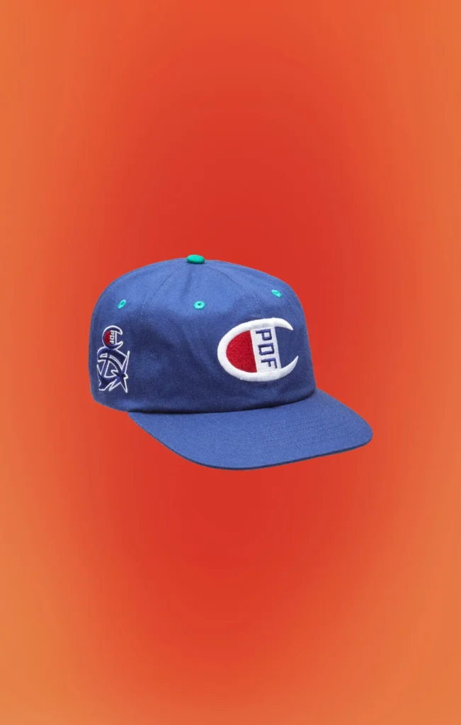 Front three-quarter view of a royal blue Champion x PDF snapback cap with oversized Champion "C" logo embroidery, white side graphics, flat brim, and green accent eyelet