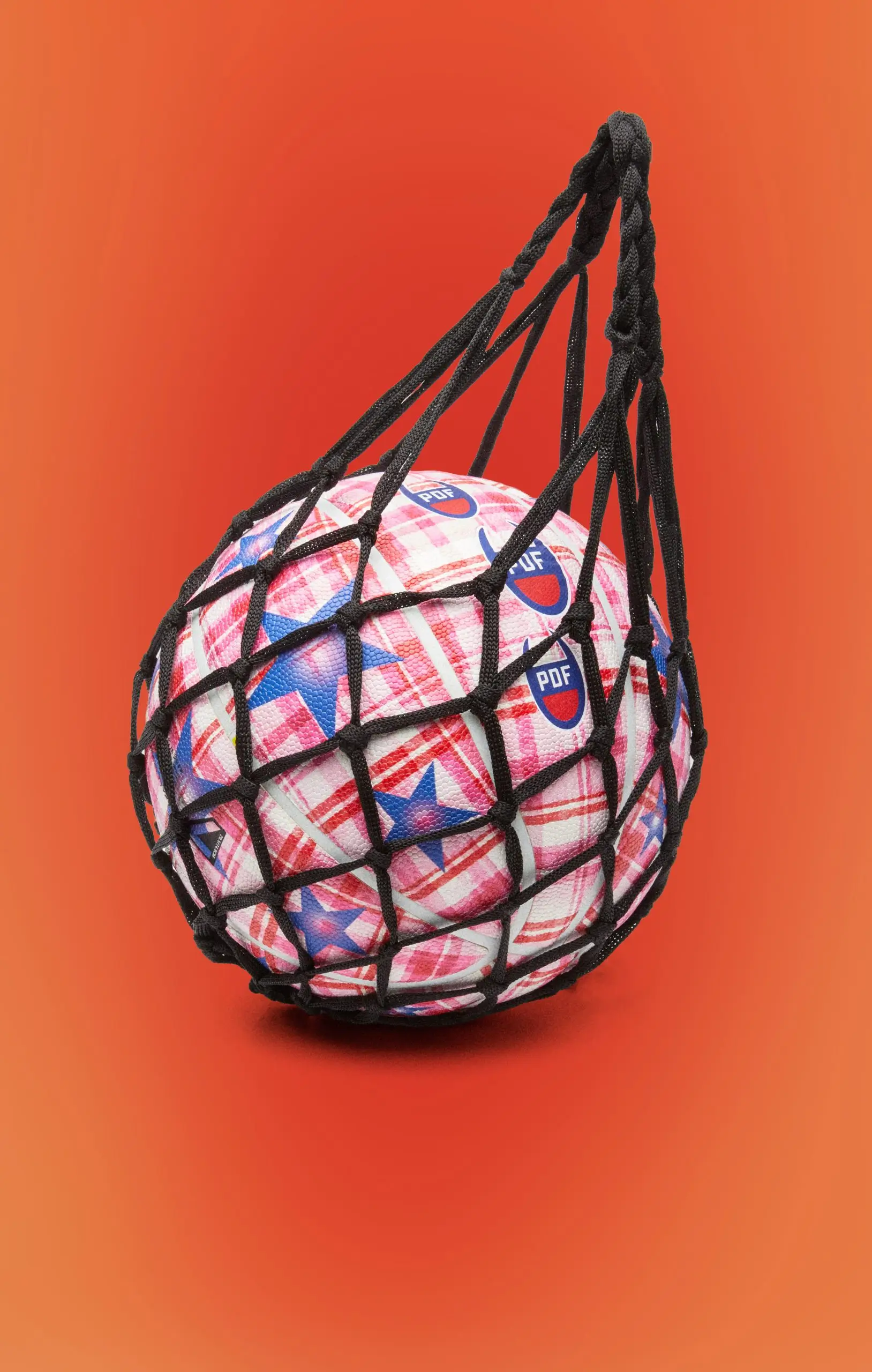 Pink plaid basketball with blue star graphics and repeating PDF logos carried inside a black rope net, combining sports equipment with fashion-inspired styling against a vibrant orange background.