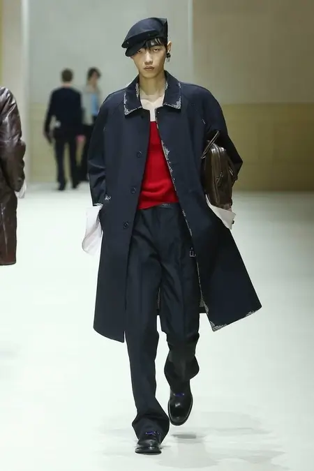 Runway model wearing a navy oversized coat over a bright red knit top, relaxed dark trousers, a black beret, and polished leather shoes while carrying a brown leather clutch during a luxury fashion show.